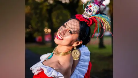 Red fascinator with skull with colourful illustrations. She is wearing an oufit with white frils, red body and a yellow/pink/yellow dress