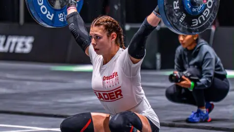 A teenager with long dark, braided hair and wearing a white shirt, shorts and knee pads, crouches down as she lifts a bar with weights on either end. 