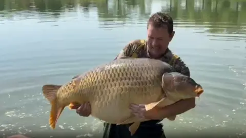 Russell Bryant A man looks down at a gigantic fish - a carp - which is grey, gold and yellow in colour. The man looks like he's struggling to hold the large fish.