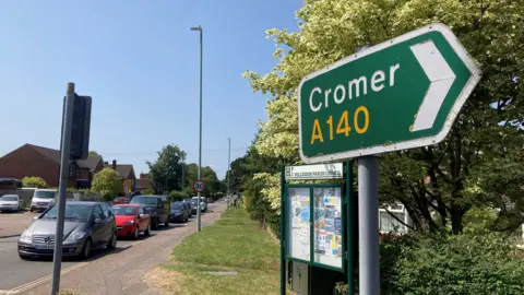 Jenny Kirk/BBC A green signpost with writing on it saying A140 to Cromer. Traffic is stationary in the background while it waits at traffic lights.