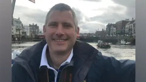 A man with short grey hair, wearing a blue shirt, brown fleece and navy jacket, sits on a boat. A canal can be seen behind him with tall buildings. 