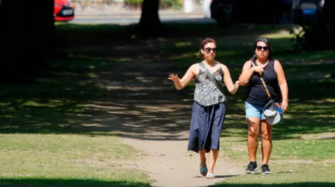 Two women walk side-by-side in Richmond Green, London as temperatures look set to soar to 38C.