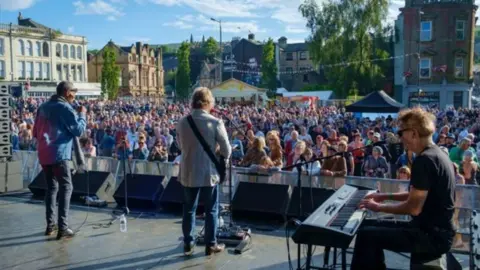 BwD Council Generic image from the back of the main stage showing a male singer, a guitarist and keyboard player watched by hundreds of people crowded before the stage in the sunshine