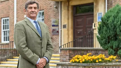 Wiltshire PCC A man with short grey hair wearing a light blue shirt, navy and white spotted tie and a tweed suit jacket standing with his hands together in front of a red-brick building with a large dark brown door and yellow flowers in a large planter. 