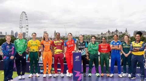 Getty Images Eleven female cricket captains, wearing their respective national team kits, stand in a line on a purple stage for a group photograph. The ICC Women’s T20 World Cup trophy sits on a plinth in the centre. The London Eye and the River Thames are visible in the background under an overcast sky.