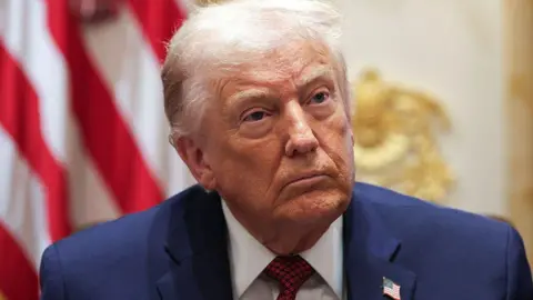 Donald Trump sits indoors wearing a dark blue suit and striped red tie, looking slightly upward with a serious expression. A blurred US flag is visible behind him.