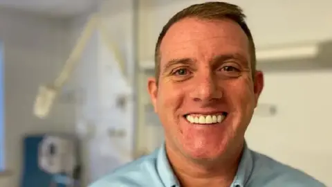 Ricky smiling looking at the camera whilst standing in a hospital cubicle. He has short brown hair and wears a blue shirt. It is a head shot of him.