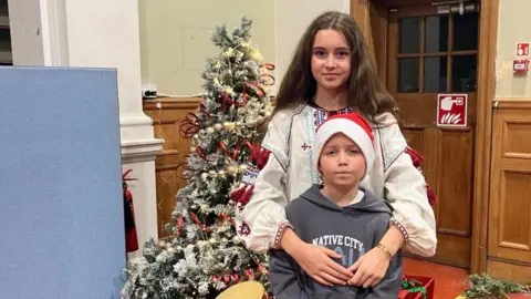A young girl with long brunette hair wearing a white jumper is standing behind her younger brother with her arms around his shoulders. The boy is wearing a grey hoodie and a Santa hat. Behind them is a Christmas tree with gifts underneath. 