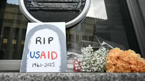 A sign placed reading "RIP USAID 1961-2925" and flowers are placed beneath the covered seal of the US Agency for International Development headquarters in Washington, DC