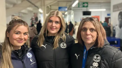 Shariqua Ahmed/BBC Three woman - Abby, Emily and Rachel, all with golden hair, wearing black Posh labelled coats, smiling for the camera.