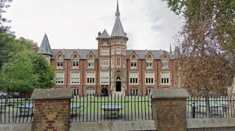 Google MVPA pictured in its large Victorian red-brick school building with tall arched windows and a central tower, viewed from behind a black metal fence, with trees and an empty courtyard in the foreground.