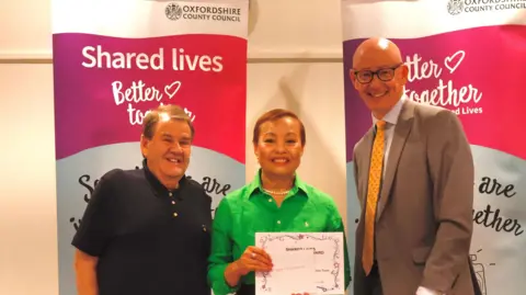 Oxfordshire County Council Two men and a woman stand together in front of a Shared Lives banner, holding up a certificate.