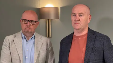 Two men standing beside each other. The man on the left is wearing glasses, a blue shirt and grey jacket. The man on the right is wearing an orange t-shirt and dark grey jacket. 