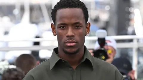 Micheal Ward, surrounded by photographers and wearing a dark green shirt and earring, looks straight into the camera