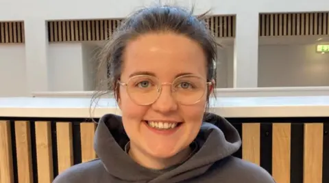 Ella Smith, with dark hair and glasses, is smiling at the camera.