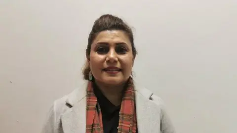 Treety Farahmand, a woman with dark hair, wearing a grey coat, a black top and a tartan style red scarf.