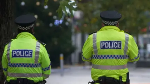 PA Media Two police officers stand with their backs to the camera. Their uniforms are florescent green jackets with the word POLICE in white block capitals on a blue background. Liverpool city centre's Lord Street can be seen in the background, out of focus