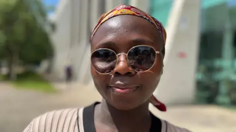 Umyima is wearing reactive glasses that have darked in the direct sunlight. She is sat outside, wearing a beige and black knitted dress and a red and yellow patterned head scarf. She has brown skin and is smiling at the camera. Behind her, slightly blurred, is a building and trees. It is a head and shoulders shot of her.