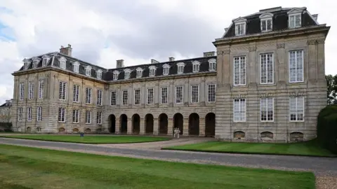 The exterior of Boughton House, near Kettering, showing a grand stone building with small archways and dozens of windows across three floors. In front is a paved driveway and lawn. 