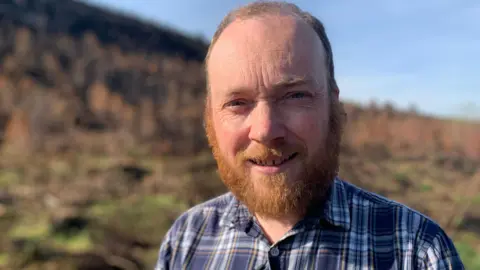 JohnRoss McMahon, he has short brown hair and a medium length ginger-brown beard. He is wearing a dark blue and pale blue checked shirt with some yellow detailing. Behind him a blurred grassy landscape can be seen.