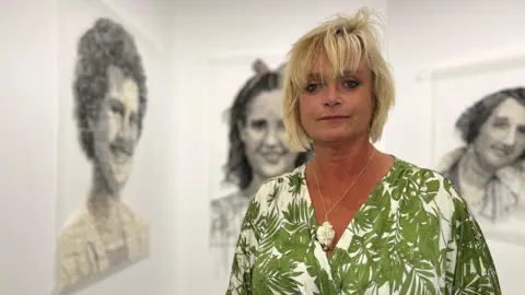 Trudie has short blonde hair and stands in an art gallery. She is wearing a green and white leaf-patterned dress and a necklace with a large pendant. Behind her, three portrait artworks are displayed on the walls, each showing different individuals.