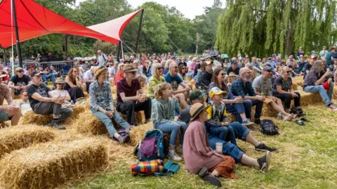 Red Rooster Dozens of festival-goers sitting down, with many of them sitting on hay bales.