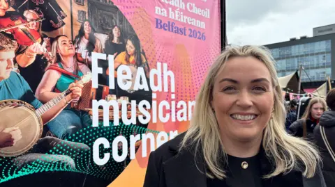 Bronagh McClean. She has long blonde hair, wearing a black blazer and black top with gold necklace. She is standing next to a banner featuring trad musicians reading 'Fleadh musician corner'.