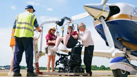 A young man is being hoisted into a light aircraft from a black wheelchair. He is on a runway alongside four adults who are providing support.