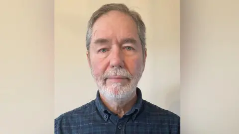 Martyn has a head of short, receding grey hair and a neatly trimmed grey beard. He is wearing a dark green and navy chequed shirt. 