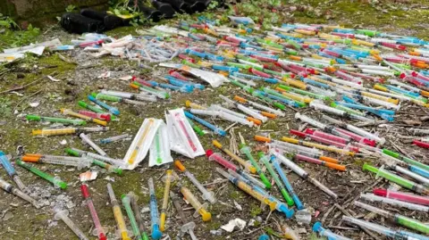 Clean Our Patch CIC Brightly coloured hypodermic needles are laid out in rows on the floor.