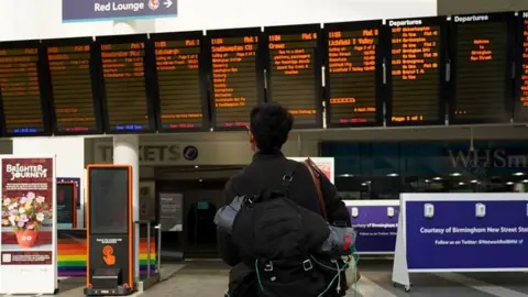 A man with a rucksack on his back has his back to us as he looks up at several train departure boards in a railway station. He has black hair and below the boards are several signs for the station.