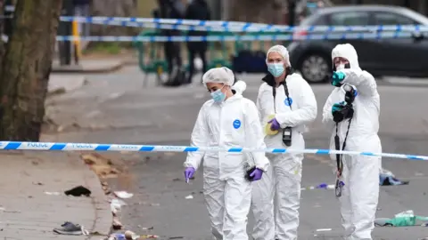 Three women dressed in white forensic suits standing behind police tape with debris on the floor