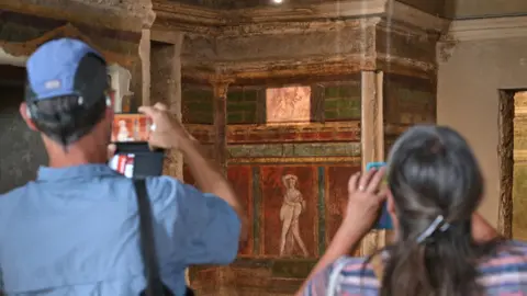 Getty Images Two tourists wearing hats visit the Hall of Mysteries in the 'Villa of Mysteries' in Pompeii. They take photos on her phone of the wall paintings, which show images of Roman life