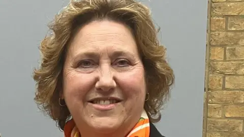 Amy Holmes/BBC A woman with wavy dark blonde hair smiles at the camera. She is wearing an orange and cream scarf.