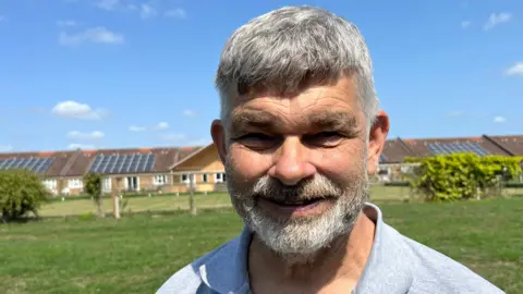 John Devine/BBC Rob Skoulding is standing off centre to the right. He has short grey hair and beard and is wearing a light blue shirt. Skoulding is standing on a field which has a row of houses on it in the backgroud.