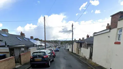 Second Street, Watling Bungalows, is a narrow road going down hill. Bungalows line either side of the street, where cars are also parked.
