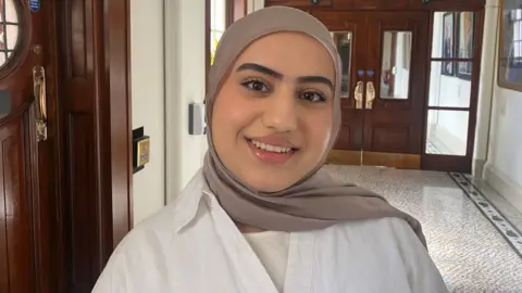 A woman standing in a corridor, she is wearing a brown hijab with a white shirt.