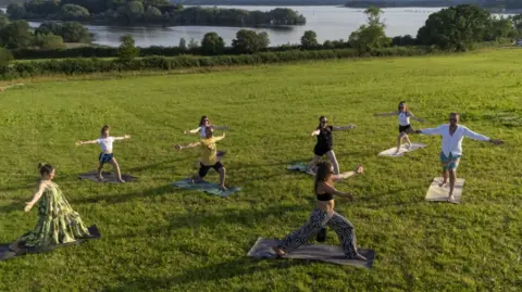 PA/Ben Birchall About six people are taking part in a yoga class in a large green field next to a lake in the background. They are all striking a pose on mats.
