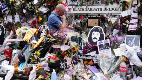 The media stood by a man surrounded by flowers, notes, and a sign that says the black seat of Saturday