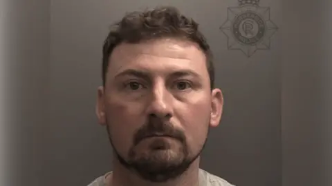 A police custody photo of Peter Coope. He has short brown hair and a short brown mustache and beard. He is wearing a grey t-shirt and is looking at the camera. The wall behind him is grey.