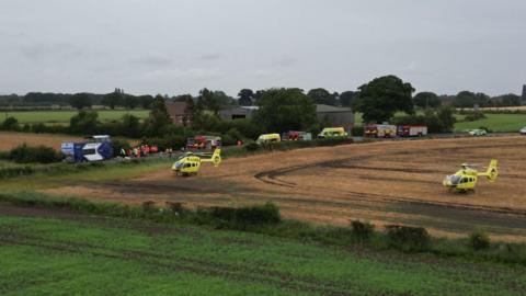 Two yellow air ambulances in a field next to a country road lined with four fire engines and two ambulances. A white and blue double decked bus and emergency workers in hi-vis jackets can also be seen