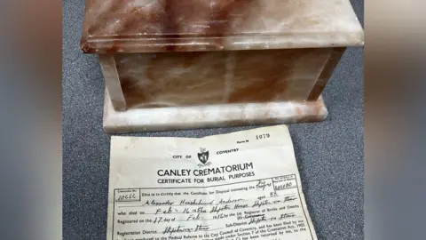 BBC A brown and white marble box and yellowed burial certificate on a grey table top. The name Alexander Anderson and age of 88 can be made out on the certificate.