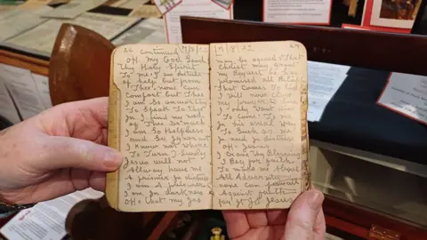 Hands are holding a small diary. The entries are written with a pencil. 