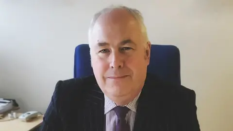 Comhairle nan Eilean Siar Malcolm Burr - a man with grey hair pictured sitting at his desk smiling wearing a pinstripe suit and purple tie