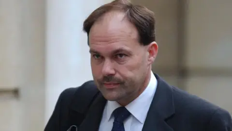 PA Glyn outside court during his 2003 trial. He has a grim look on his face, and has short brown hair and a moustache. He wears a black suit jacket and a dark tie, with a white shirt. 