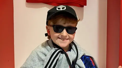 A young boy sits in a football changing room smiling at the camera. He has both thumbs raised up. He wears a black cap and sunglasses and has a grey jumper tied around his neck.