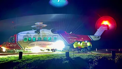 The picture shows a Coastguard rescue helicopter on the ground at night with its lights blazing so brightly that they spill across the grass around it. The rotors are spinning, creating a blurred circular halo above the aircraft. The helicopter’s red and white body stands out against the darkness, and the tail light glows with a bright red ring.