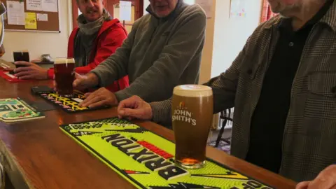 The picture shows men drinking half a liter of beer in a pub bar. "John Smith's" beer glass in the foreground. 