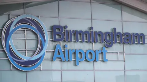 Reuters The Birmingham Airport sign on the side of a building with Birmingham in dark blue letters and airport in light blue letters. The airport's circular symbol is to the left of the words.