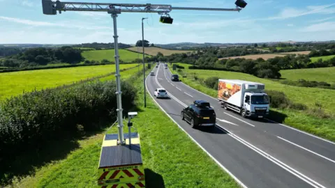 Devon County Council A high angle view of the road showing it flowing into the Devon countryside. There are fields and trees. There are various vehicles traveling on the road, including cars and trucks. In the foreground is the AI ​​camera installed on top of a vehicle. It resembles a small crane attached to the top of the vehicle with cameras attached. 